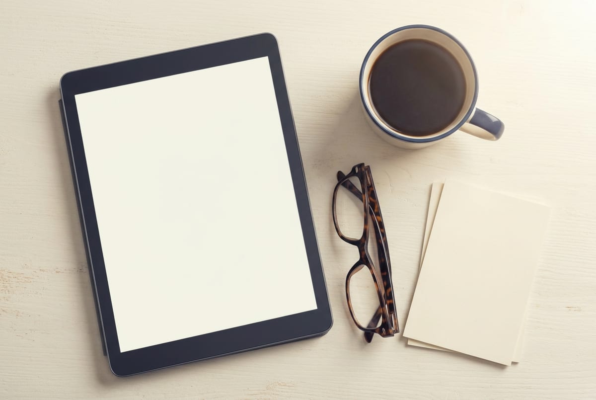 Vista superior de um tablet apoiado em uma mesa clara ao lado de uma xícara de café, um par de óculos e uma pilha de papéis.